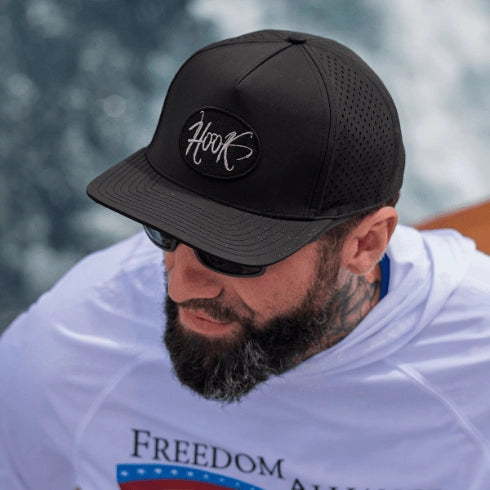 A close-up lifestyle shot of a man wearing a black hook optics trucker baseball cap with a black oval patch featuring the white logo 'Hook'. He is also wearing a white t-shirt with a patriotic graphic and hook optics billfisher sunglasses, set against a blurred outdoor background.