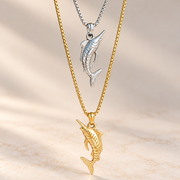 Two layered fish necklaces displayed on a light beige marble surface with soft shadows. The top necklace features a silver-toned box chain and a silver fish pendant.