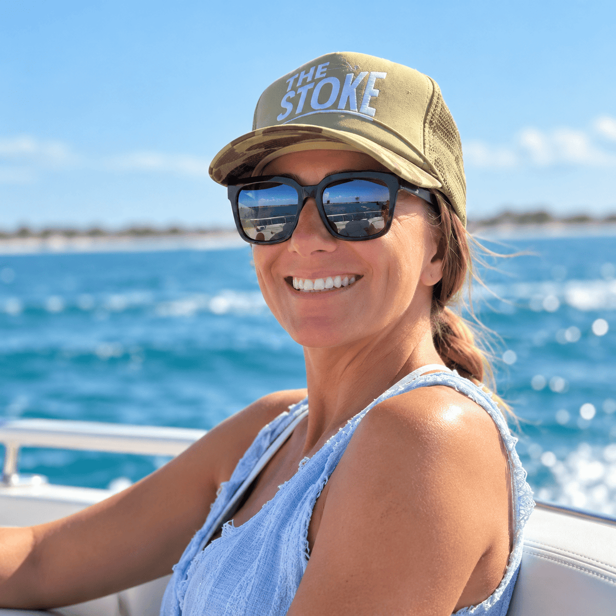 A smiling woman on a boat wearing a tan trucker hat with 'THE STOKE' in white text, black Hook Optics sunglasses, and a light blue sleeveless top, with blue ocean water in the background.