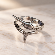 A sterling silver ring shaped like a marlin fish, resting on a light beige marble surface.