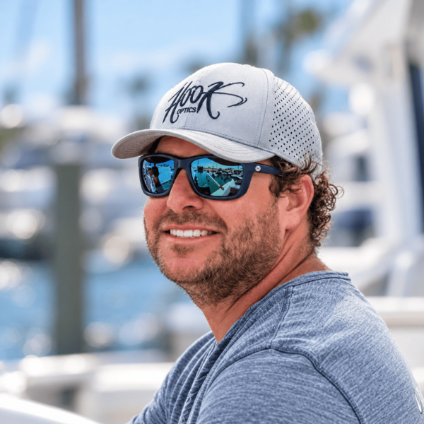 A smiling man wearing a white and gray mesh baseball cap with a black script logo that reads 'Hook Optics' and dark mirrored sunglasses, photographed outdoors with a blurred marina or boat background.