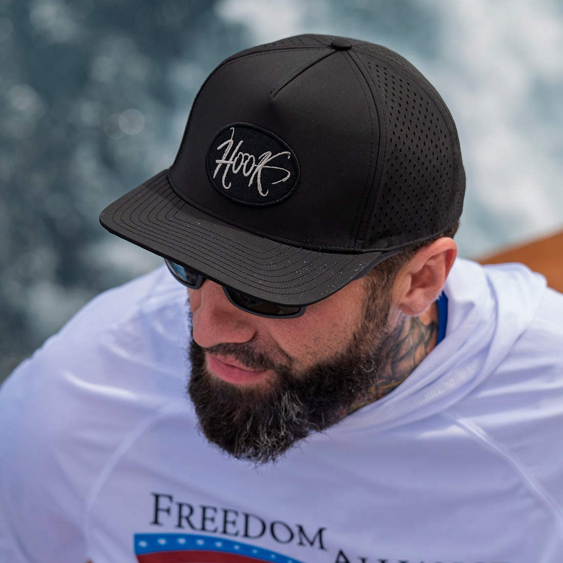 Man wearing a black cap with 'Hook' logo and sunglasses, sitting by water.