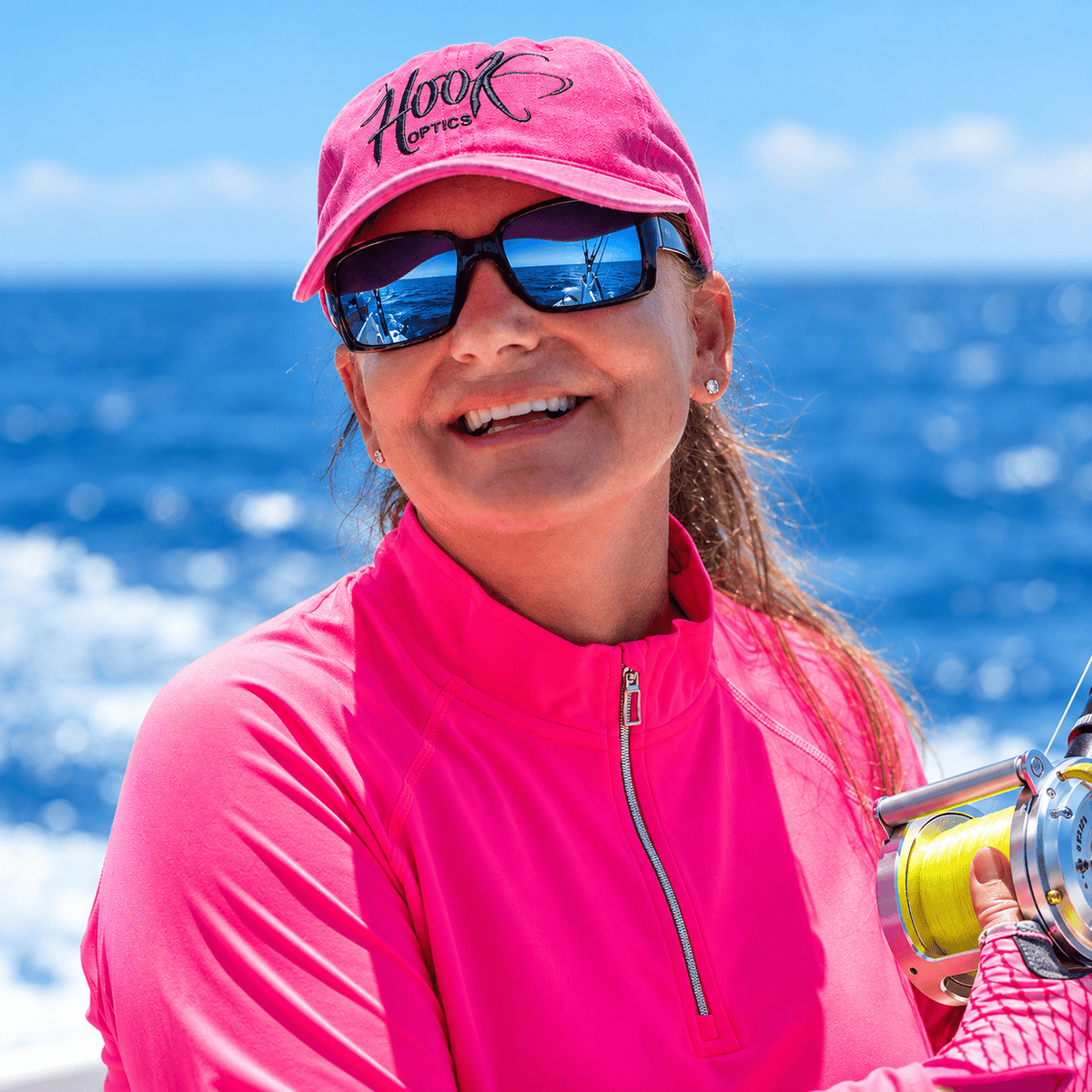 A smiling woman outdoors by the ocean wearing a bright pink cap with black script logo, mirrored  hook optics sunglasses, and a matching pink zip-front athletic top, while holding a fishing reel.