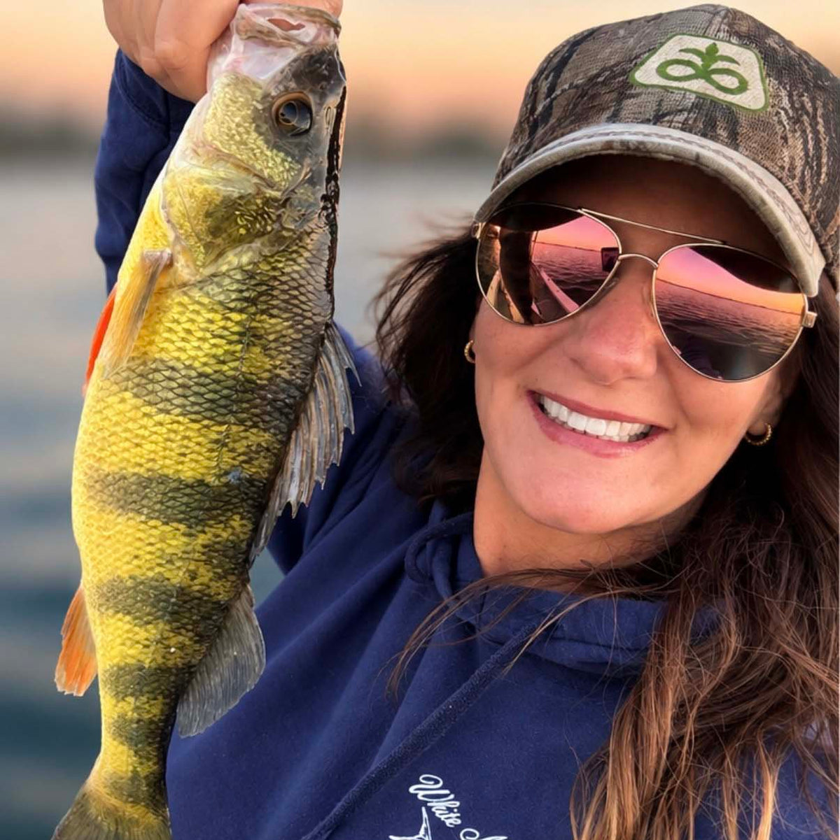 A smiling lady angler wearing a camouflage cap with a green logo and Hook Optics aviator sunglasses holds up a yellow perch fish during a fishing trip on the water at sunset.