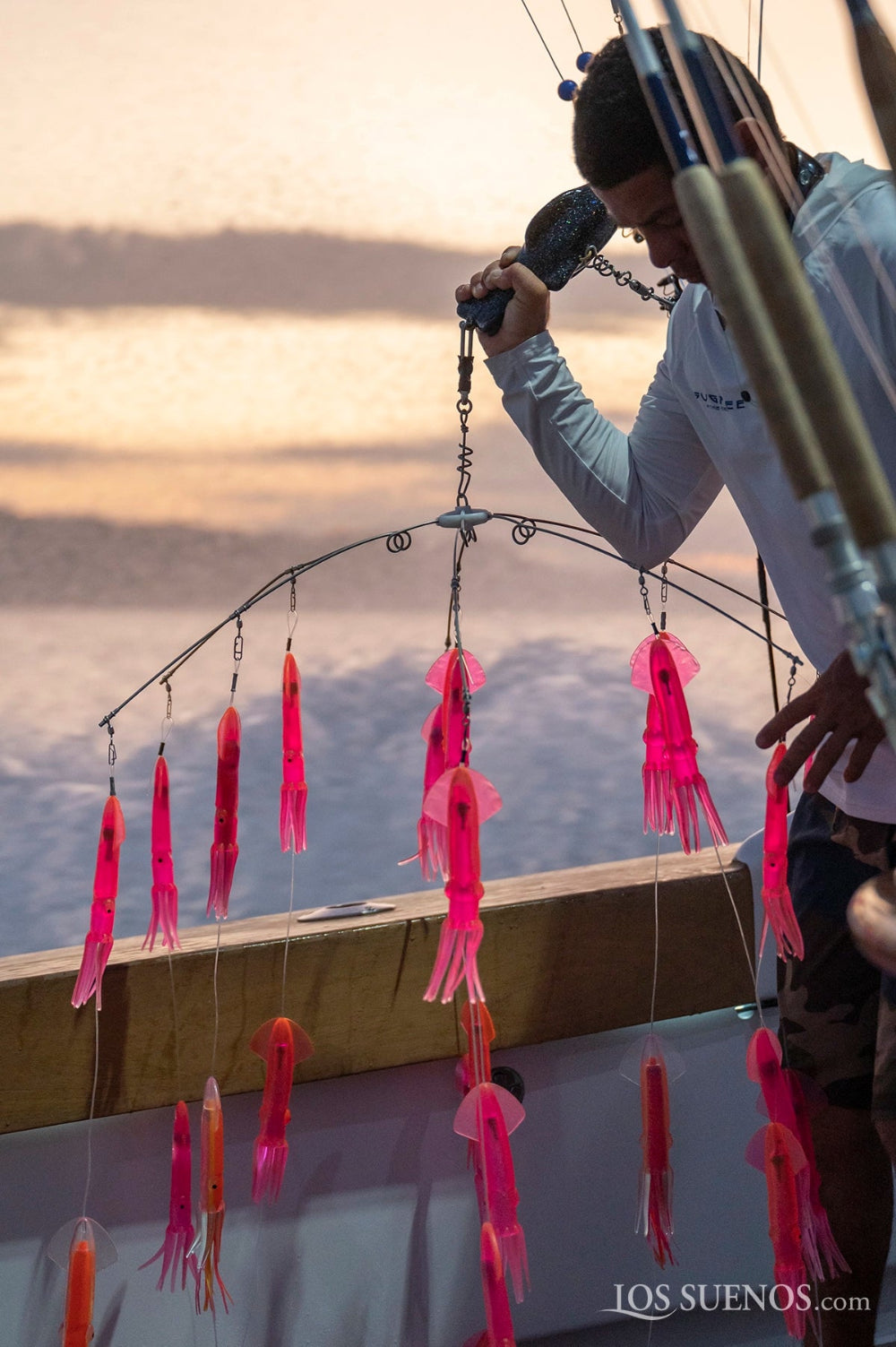 Person on a boat with pink and orange fishing lures hanging from a line against a sunset sky wearing Hook Optics Sunglasses