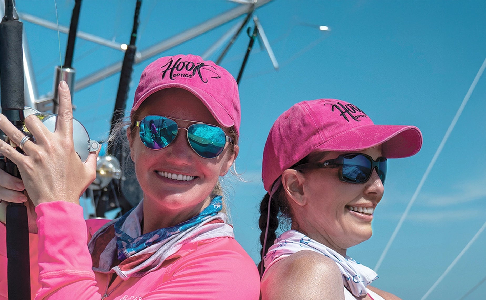 Two women wearing pink hats and hook optics best sunglasses on a boat with a clear blue sky.